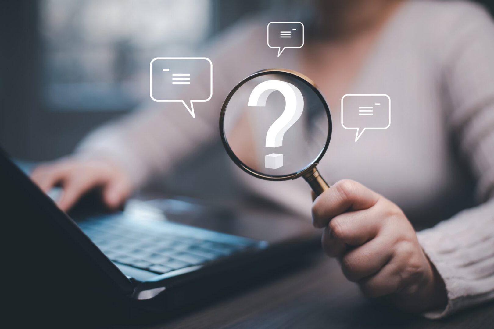 Person holding magnifying glass over question mark near laptop.